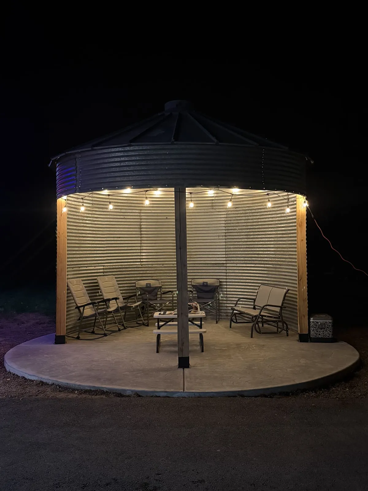 The finished grain bin pavilion at night, open-sided and lit with string lights over patio furniture